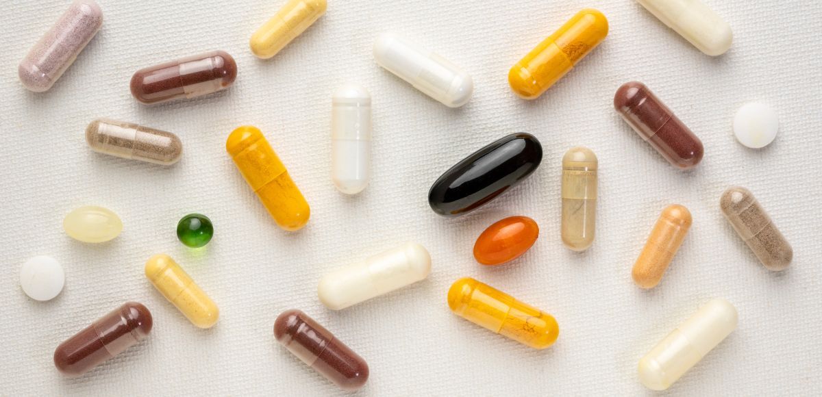 Assorted capsules and tablets scattered on a white surface, including white, yellow, brown, black, and green pills