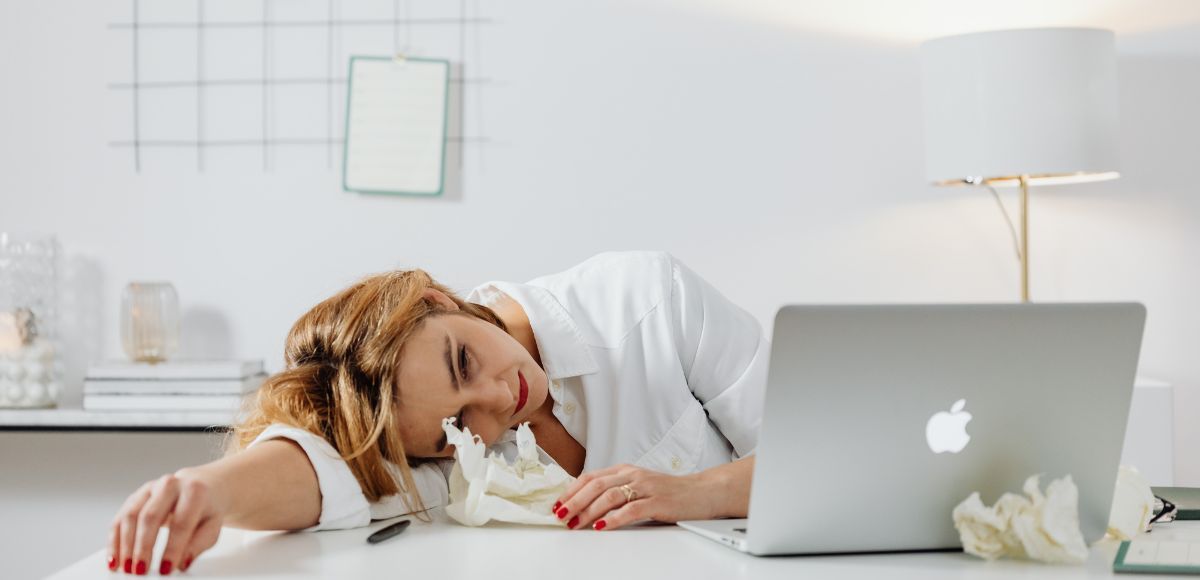 Tired person resting on a desk beside a silver laptop in a bright office