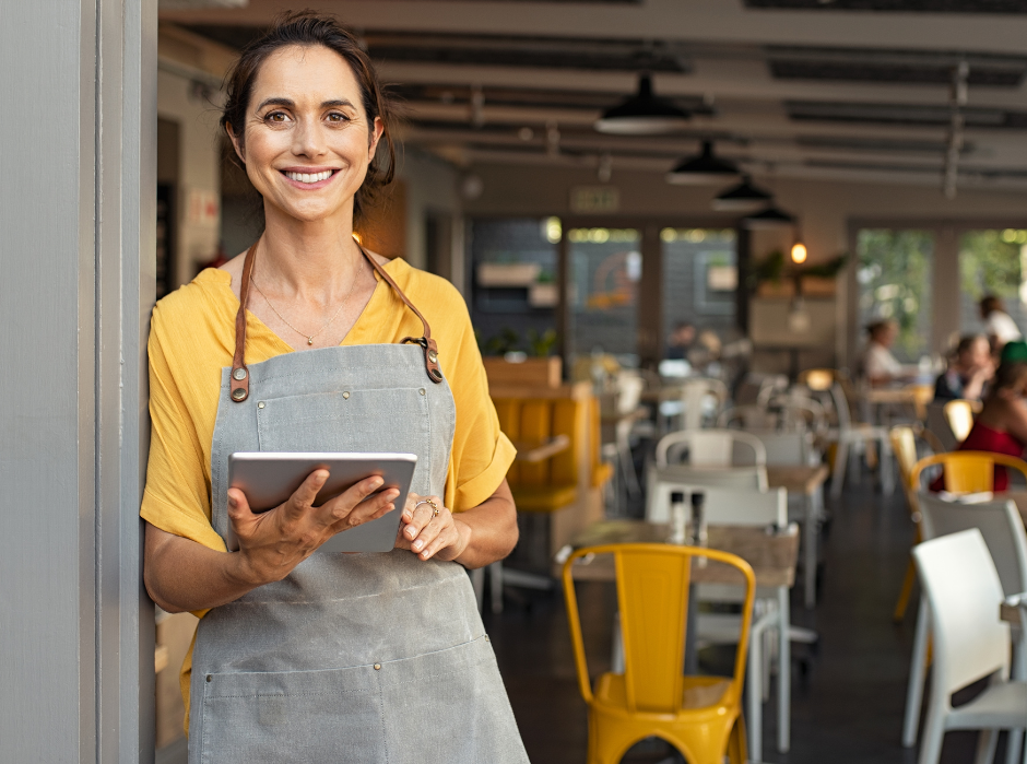 A person smiling, holding a tablet in a bright cafe setting