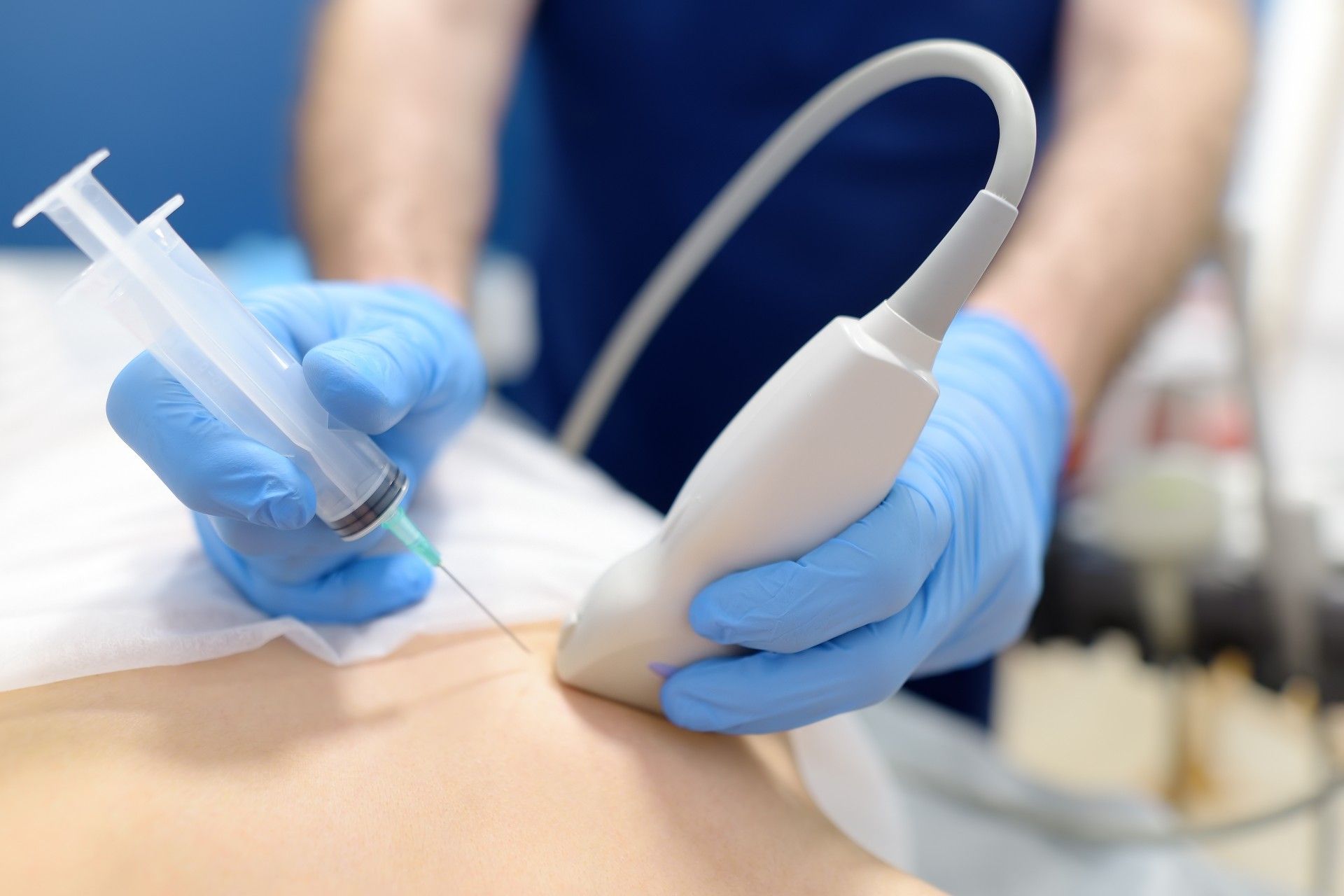 Gloved hands using ultrasound probe and syringe on a patient’s abdomen during a medical procedure