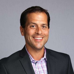 Smiling man in a dark blazer over a checkered shirt against a gray background