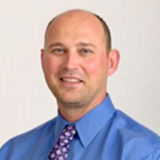 Portrait of a smiling man in a blue shirt and patterned tie against a white background