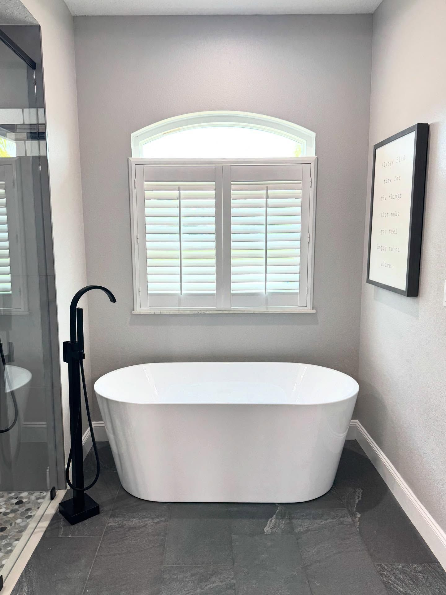 Modern bathroom with freestanding white tub, black faucet, and arched window with shutters