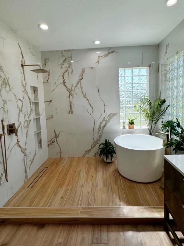 Modern bathroom with marble walls, wood floor, freestanding tub, glass shower, and potted plant by a window