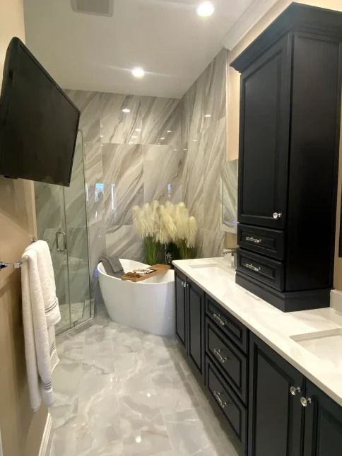 Narrow modern bathroom with black vanity, wall TV, freestanding tub, and marble-pattern tile walls
