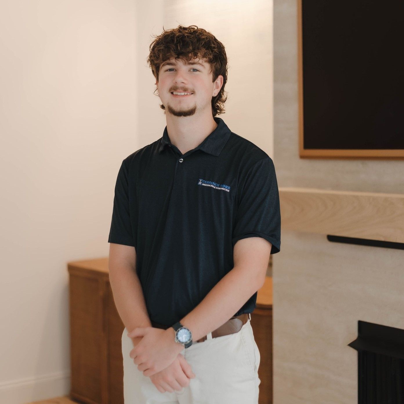 Standing indoors by a fireplace, a man in a dark polo shirt and light pants smiles with hands clasped.
