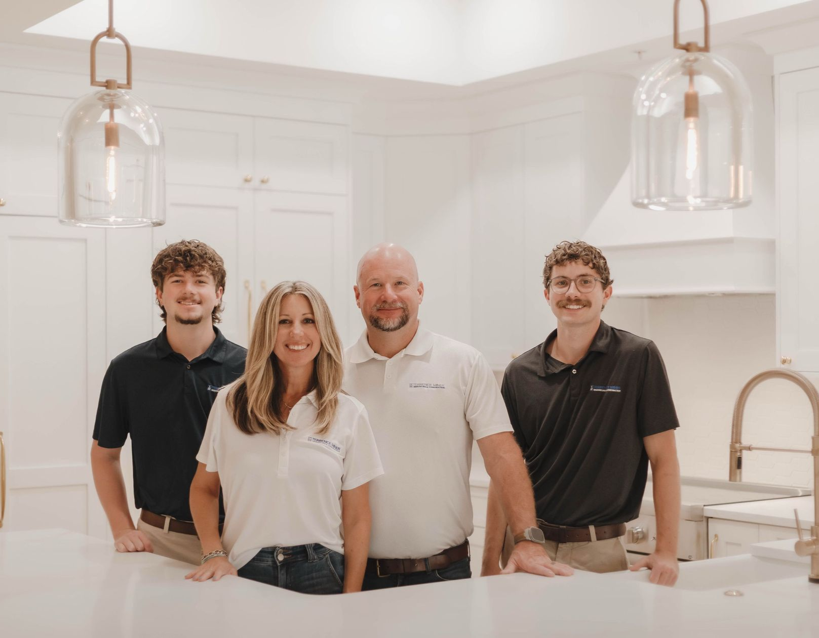 Four people standing in a bright white kitchen, smiling behind a marble island