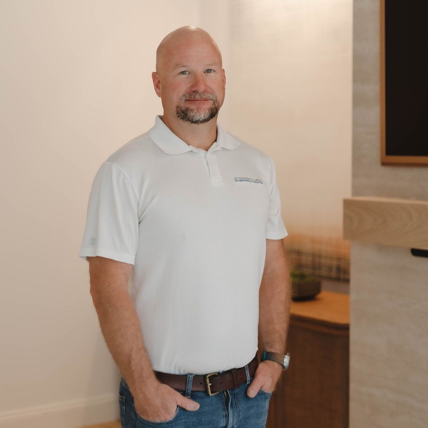 Man standing indoors in a white polo shirt and jeans, smiling near a reception desk