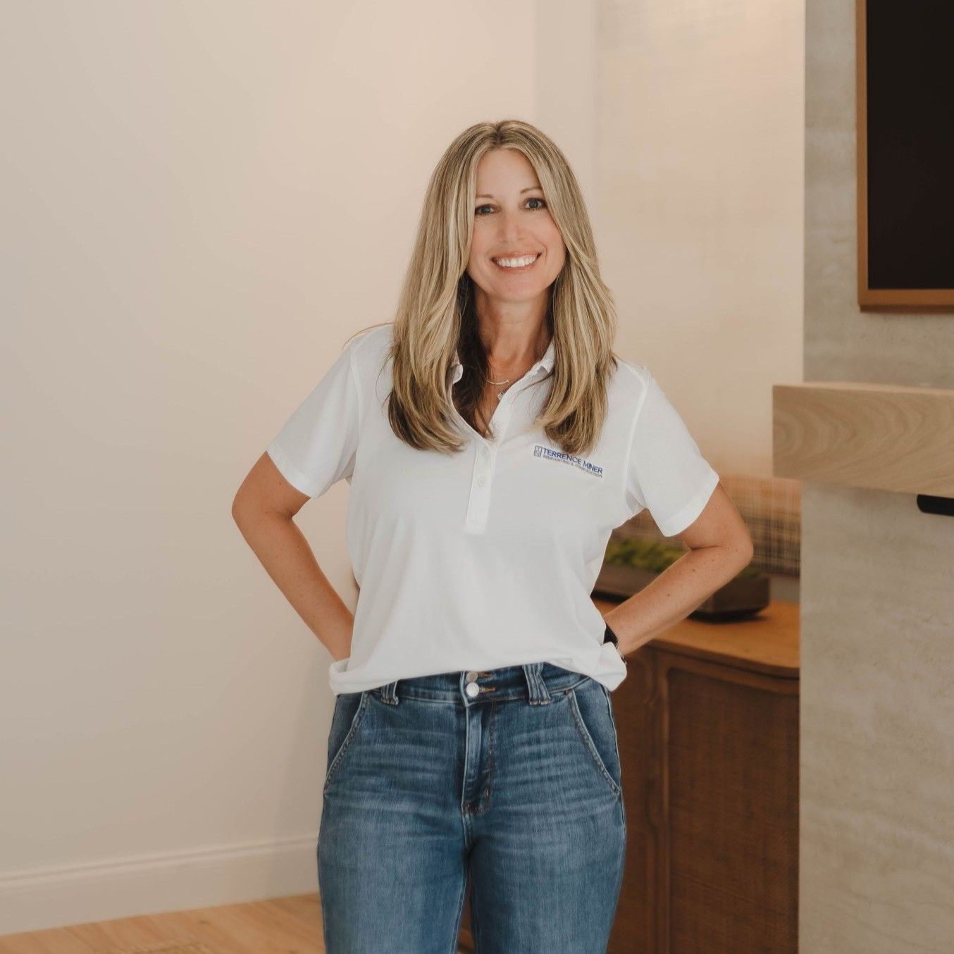 Woman in a white polo and jeans standing indoors with hands on hips and smiling