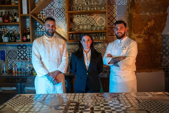 Tre membri del personale sono dietro un bancone piastrellato in un ristorante rustico, vestiti con uniformi bianche e abiti neri.