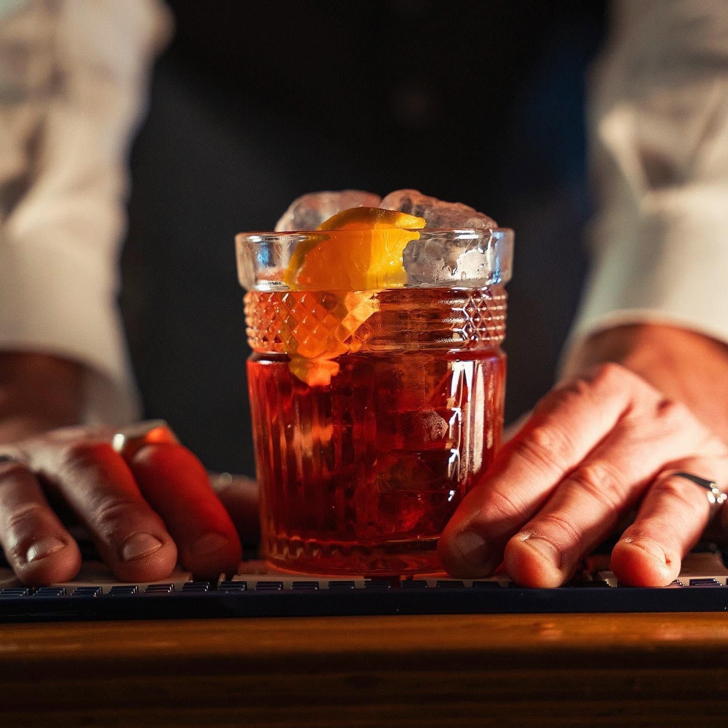 Mani appoggiate accanto a un bicchiere di cocktail ambrato guarnito con una scorza d’arancia sul bancone di un bar