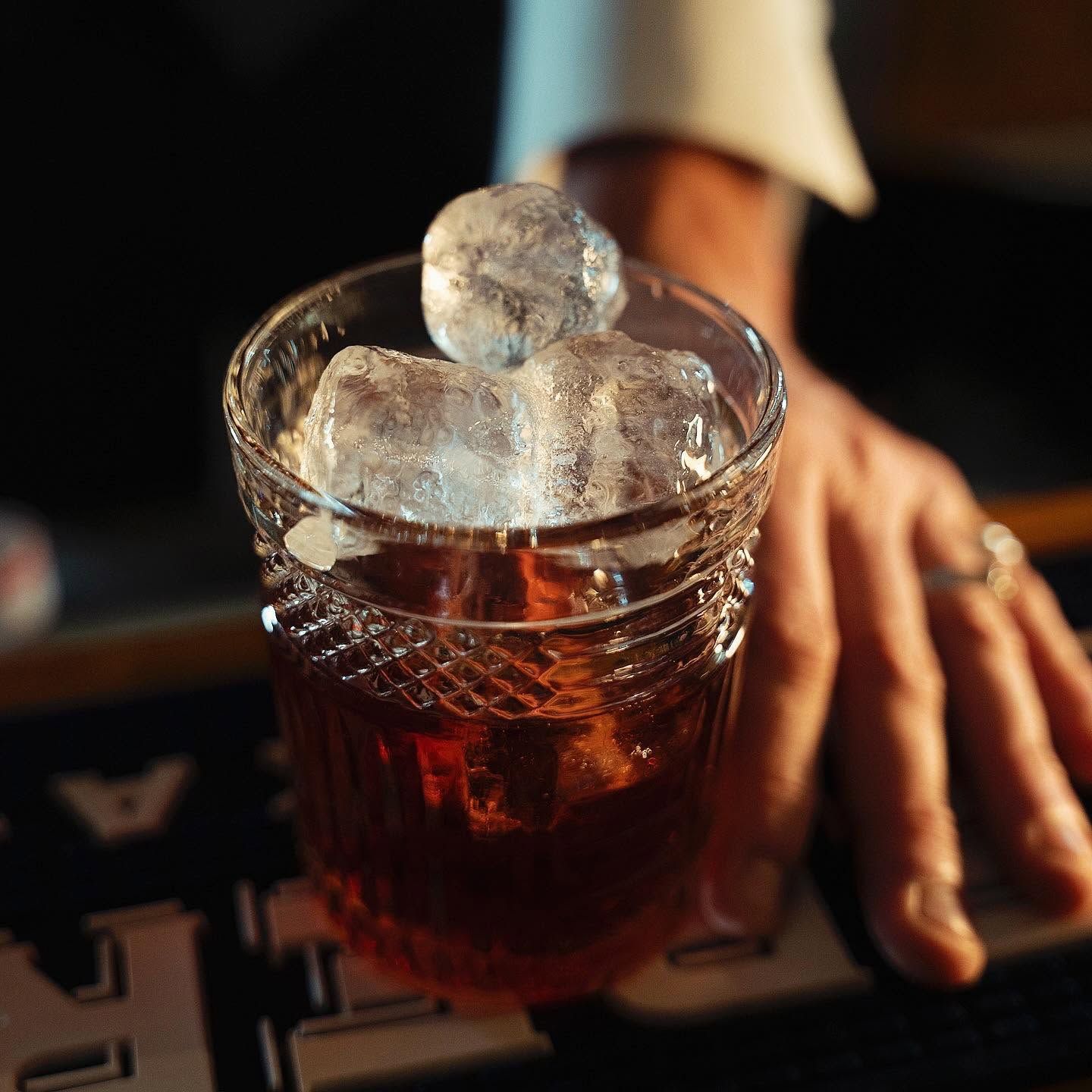 Mano che regge un bicchiere di una bevanda scura con ghiaccio sopra un mixer da DJ, in penombra