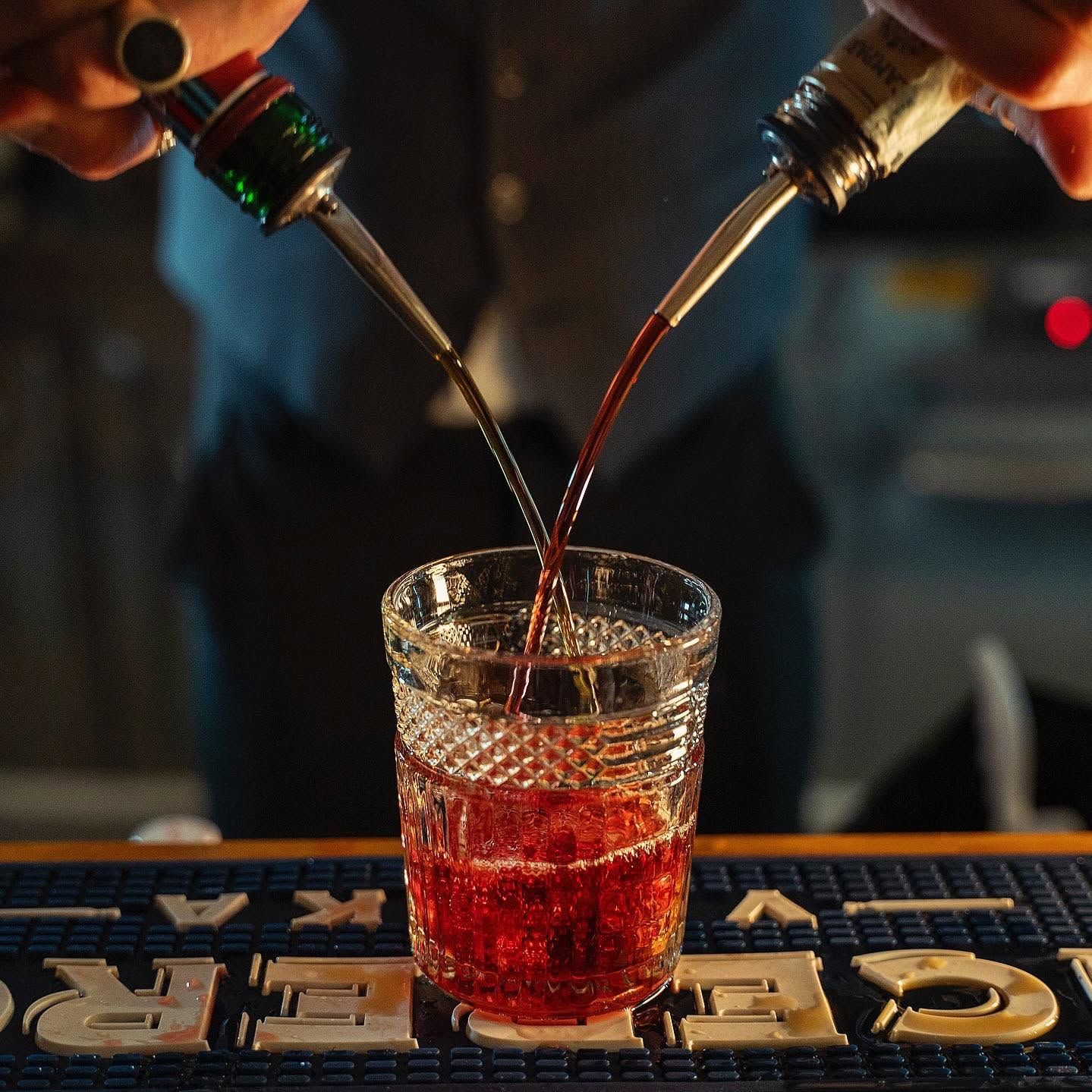 Due bottiglie versano un liquore rosso in un bicchiere su un tappetino da bar