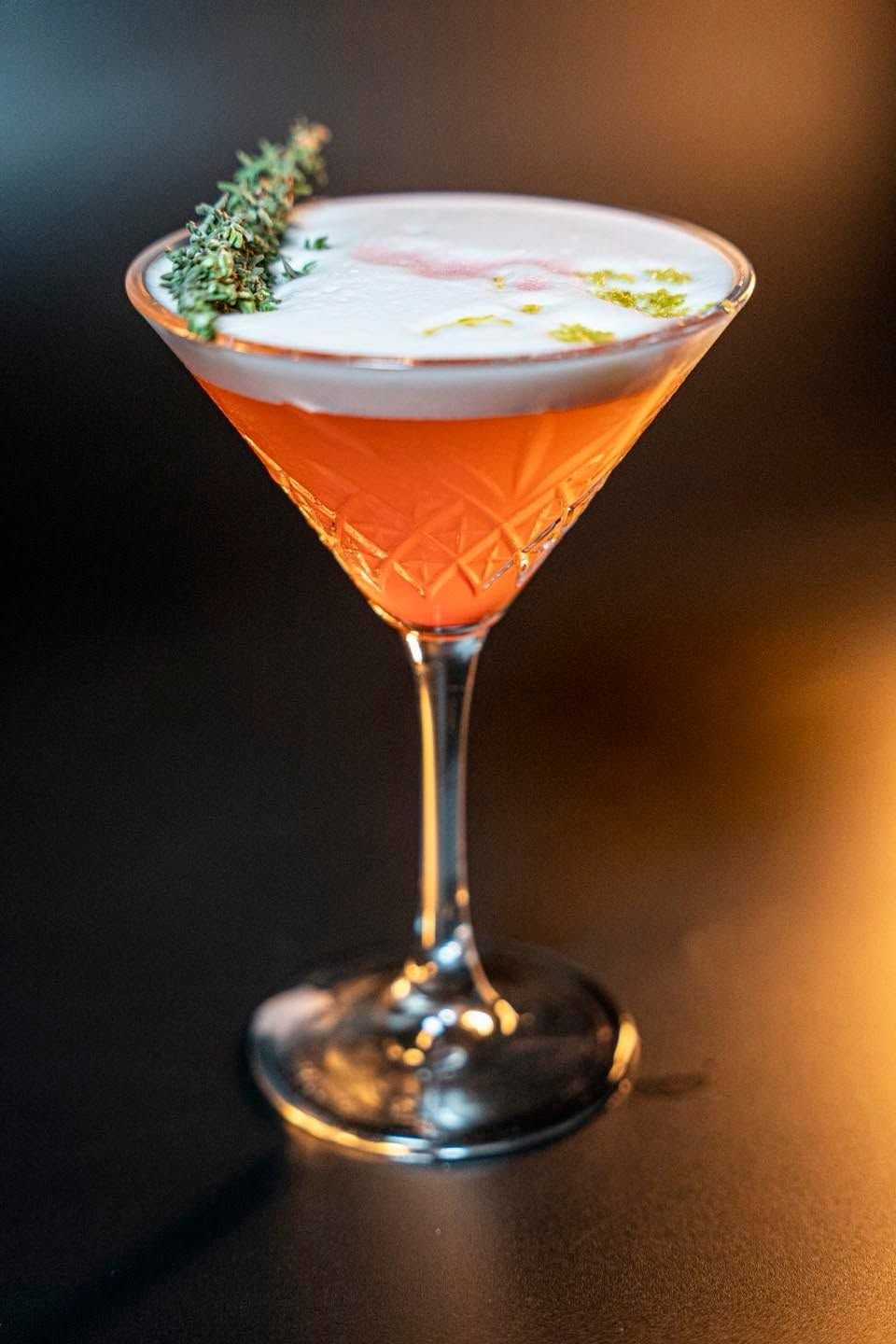 Cocktail arancione in una coppa da martini, con una schiuma bianca e soffice e guarnito con erbe aromatiche, su un tavolo scuro