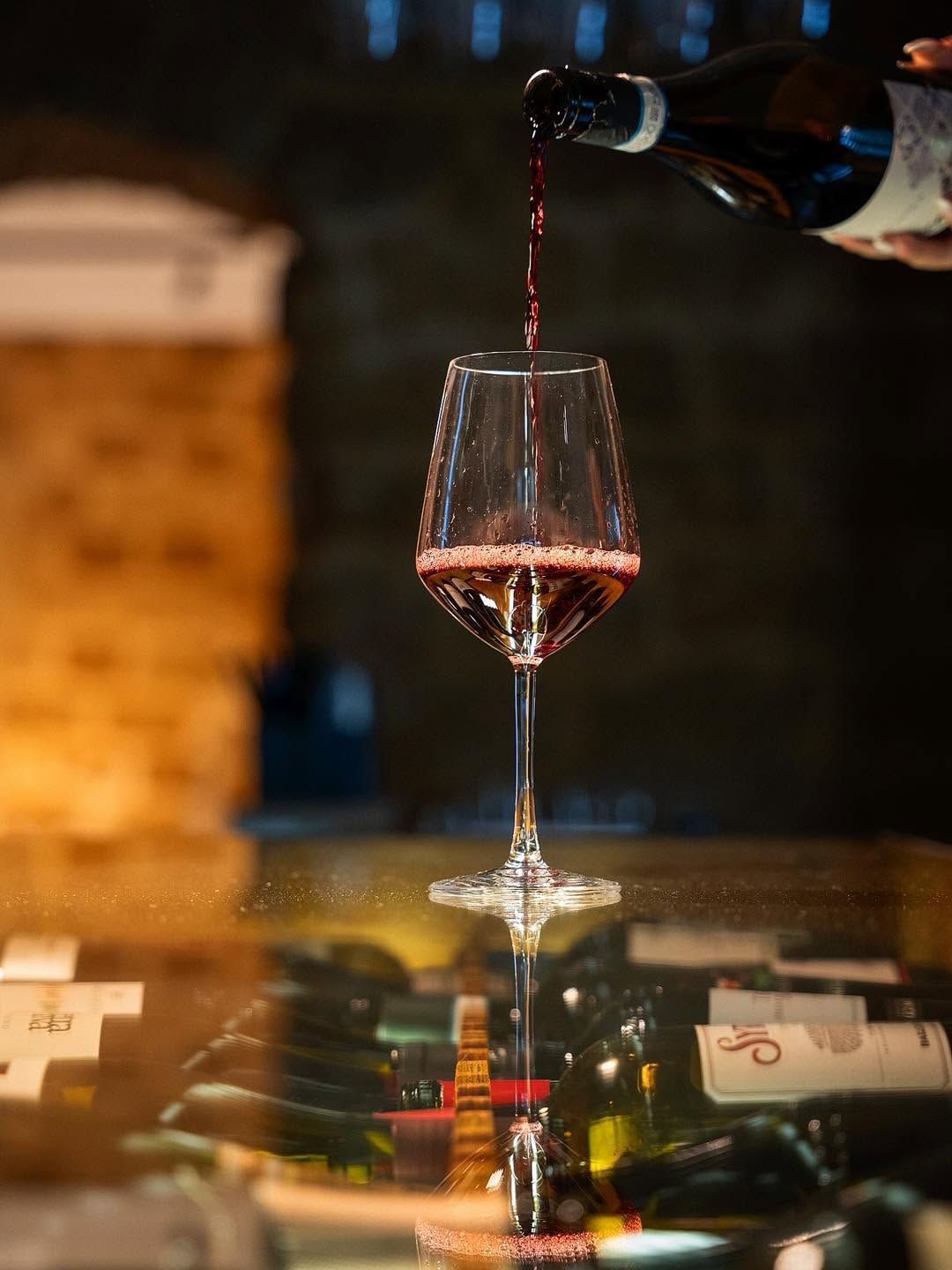 Vino versato in un bicchiere su un tavolo in penombra, con bottiglie e candele sullo sfondo