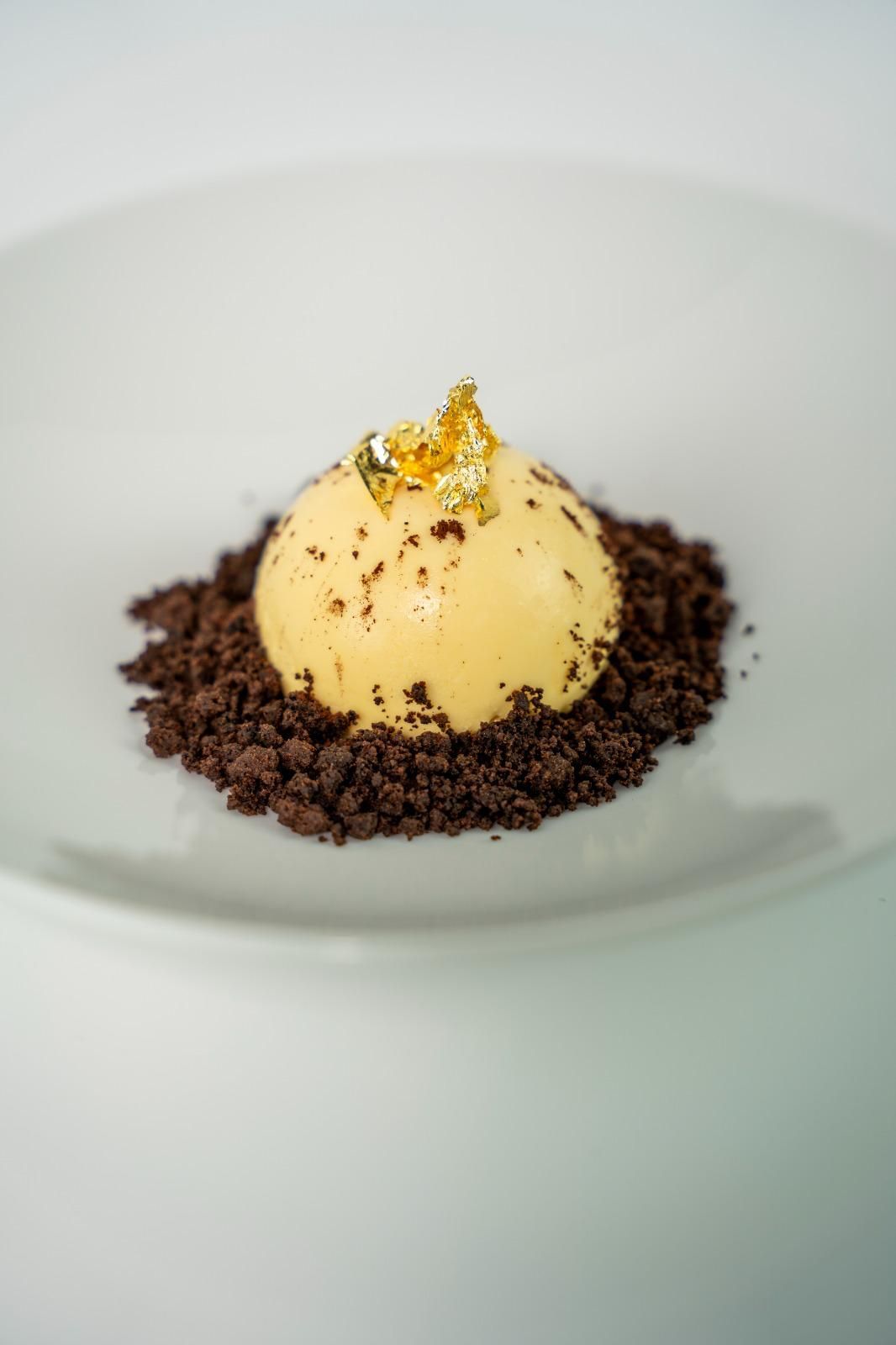 Sfera di dessert alla vaniglia su briciole di cioccolato, decorata con foglia d’oro, su un piatto bianco
