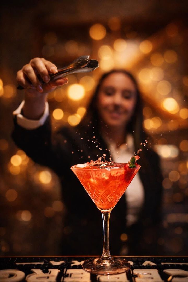 Un bartender cosparge una guarnizione su un cocktail rosso in una coppa da martini, con luci calde e sfocate sullo sfondo.