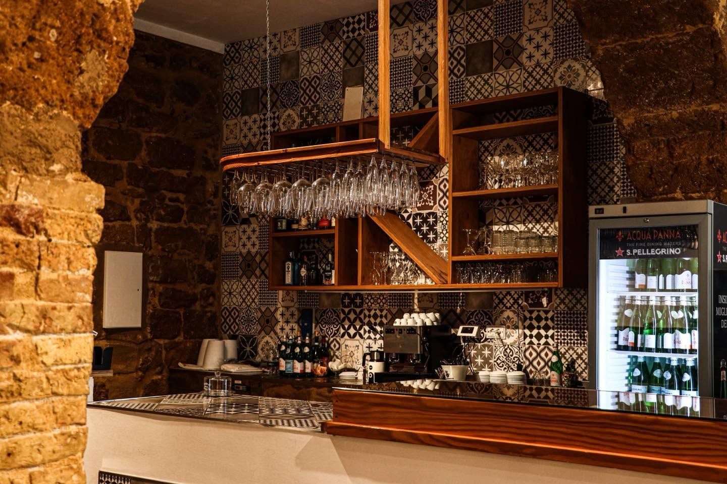 Accogliente bar rustico con scaffali in legno, parete in pietra, decorazioni con motivi e un frigorifero a vetri pieno di bottiglie
