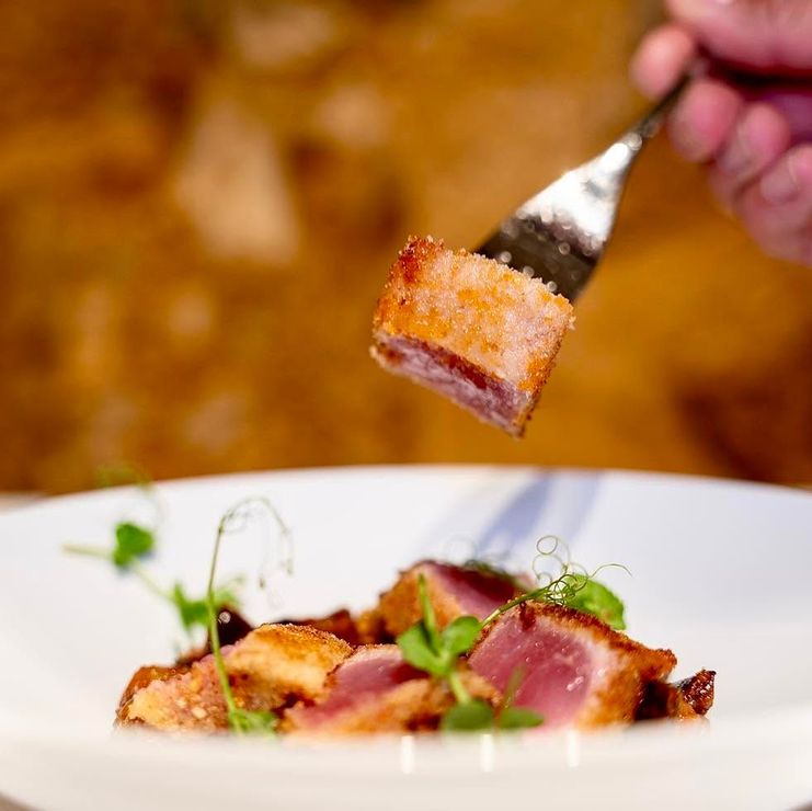 Forchetta che solleva un cubetto rosolato sopra un piatto guarnito con erbe aromatiche e fette rosa