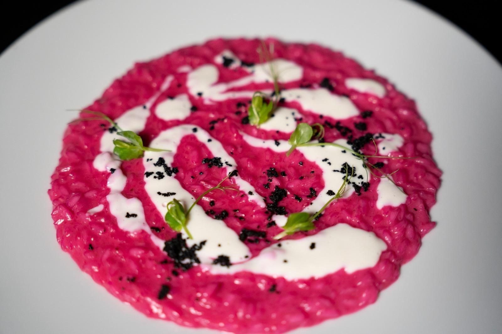 Risotto magenta guarnito con crema bianca, erbe verdi e condimento nero, servito su un piatto bianco