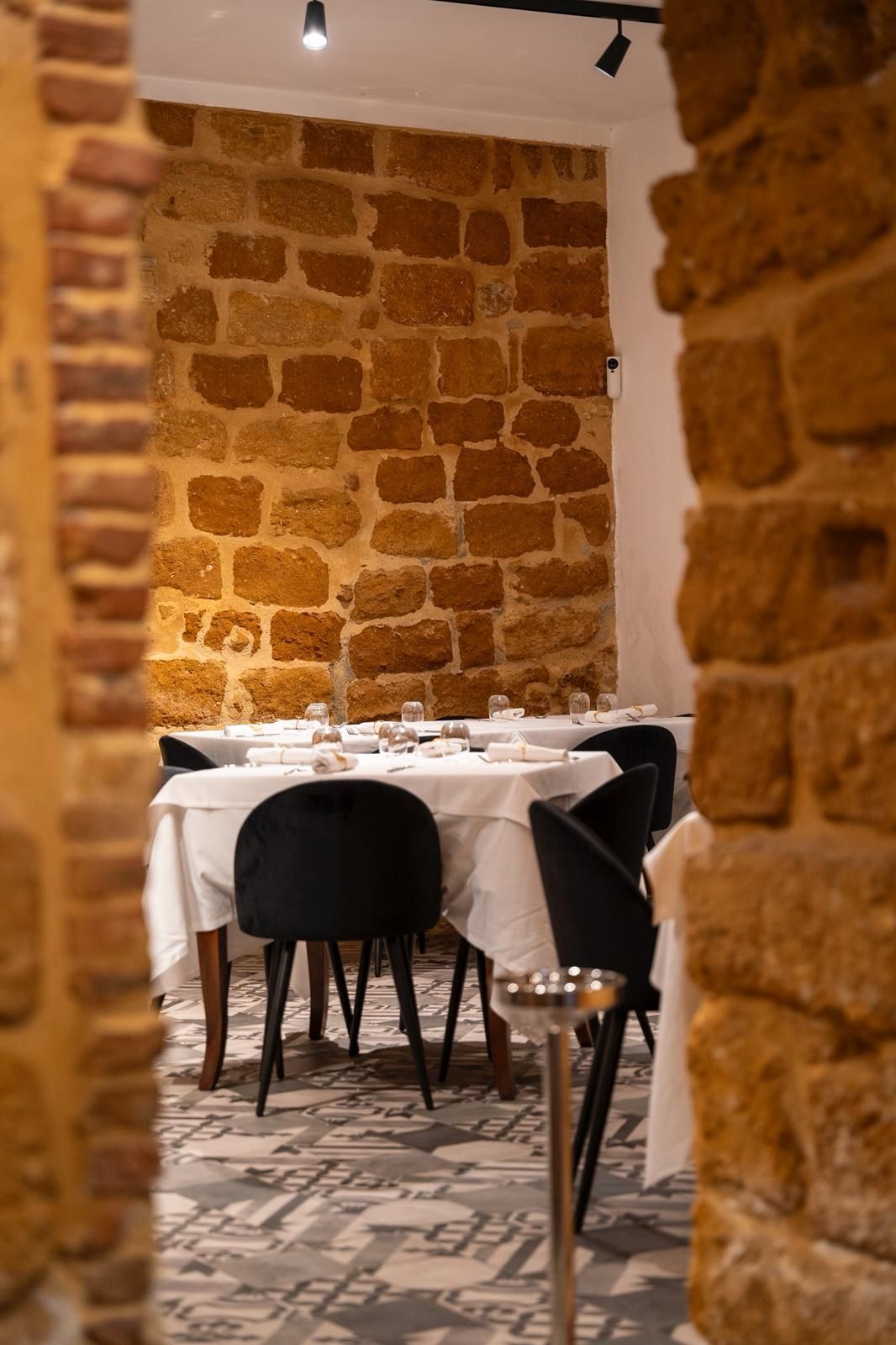 Sala da pranzo vuota di un ristorante, con tavoli coperti da tovaglie bianche, sedie nere e pareti in pietra