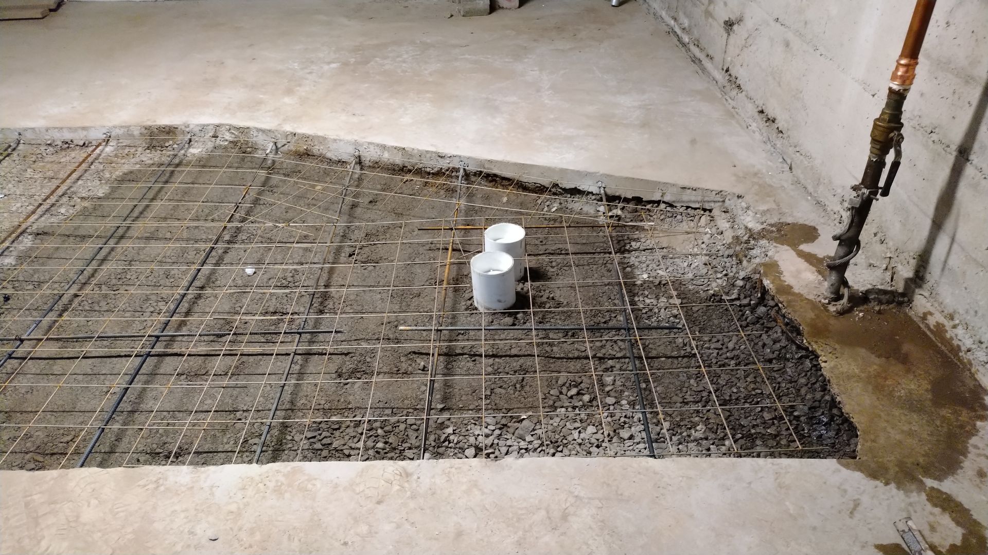 Plancher de sous-sol en construction. Treillis d'armature, deux tuyaux blancs, béton et eau à côté d'un mur en béton.