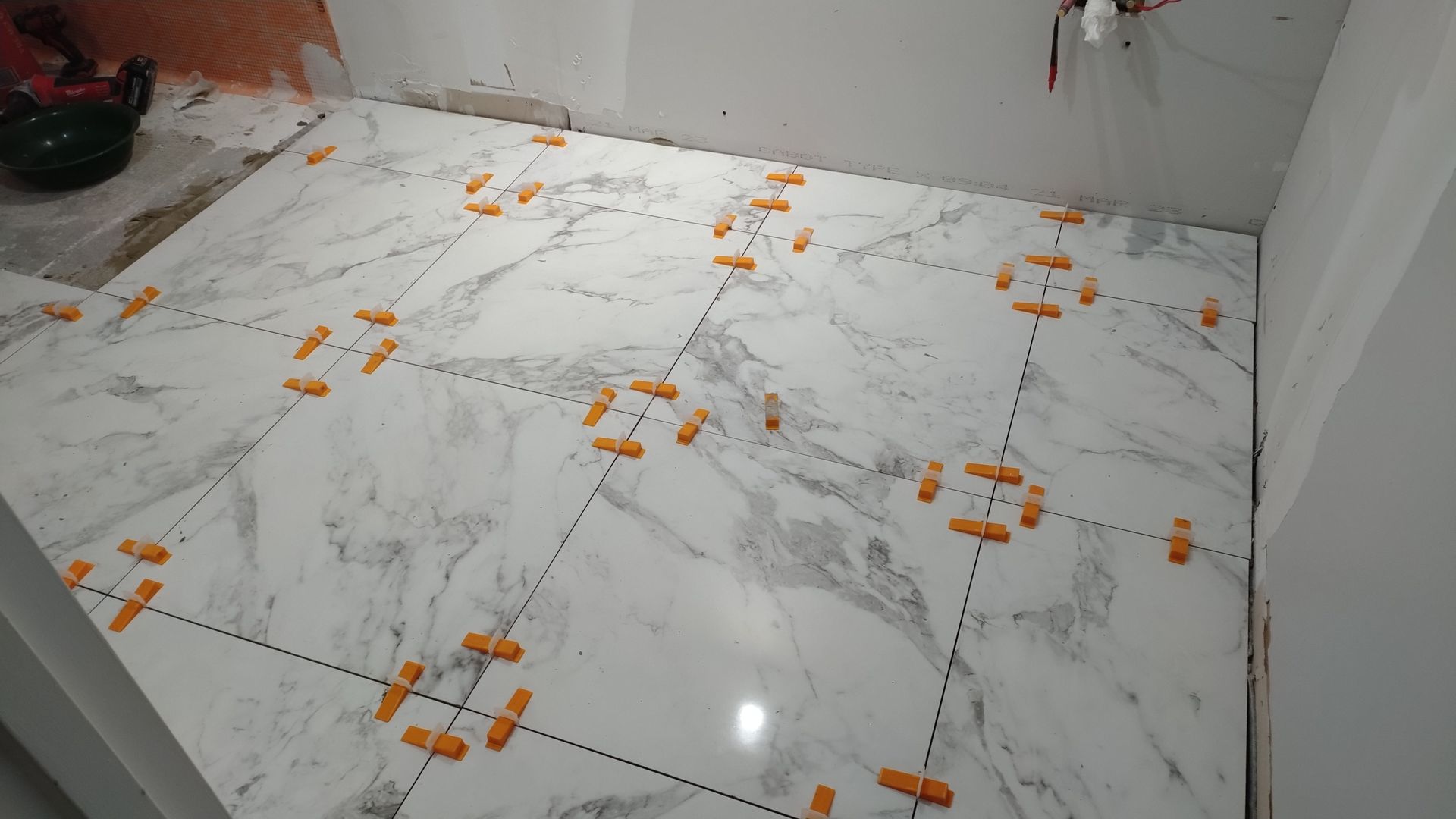 Pose de grands carreaux blancs à motif marbre au sol de la salle de bains ; clips de nivellement orange visibles.