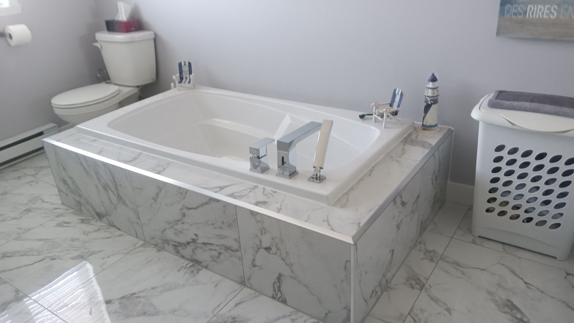 Salle de bains avec baignoire blanche entourée de faux marbre, à côté des toilettes et du panier à linge.