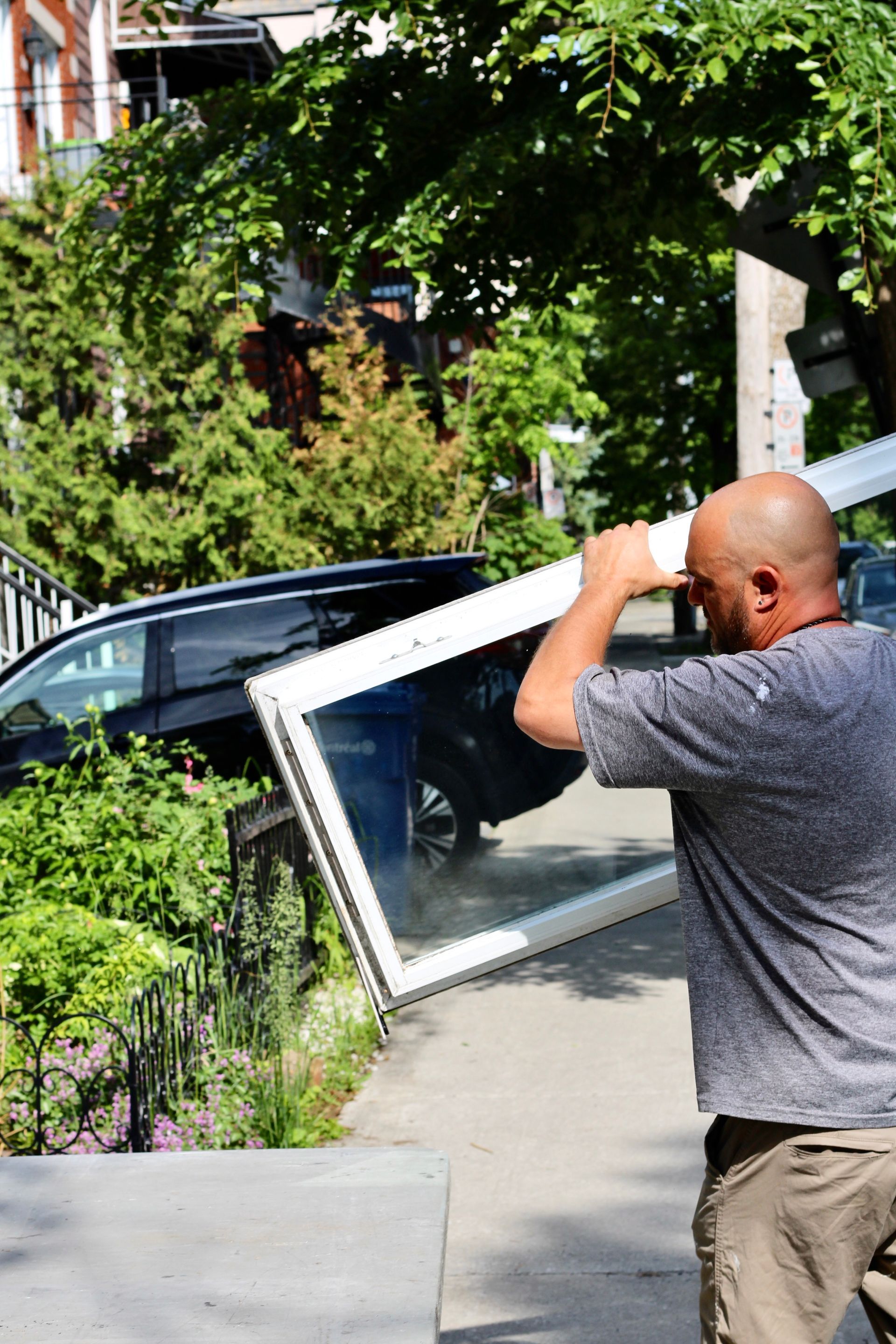 Man carrying a framed window through a sunny front yard beside a parked car