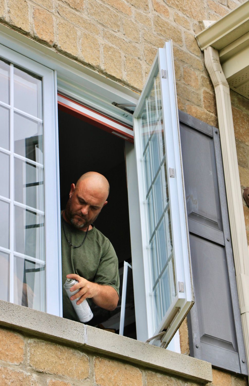 Man leaning out of a second-story window, using a tool on the white window frame.