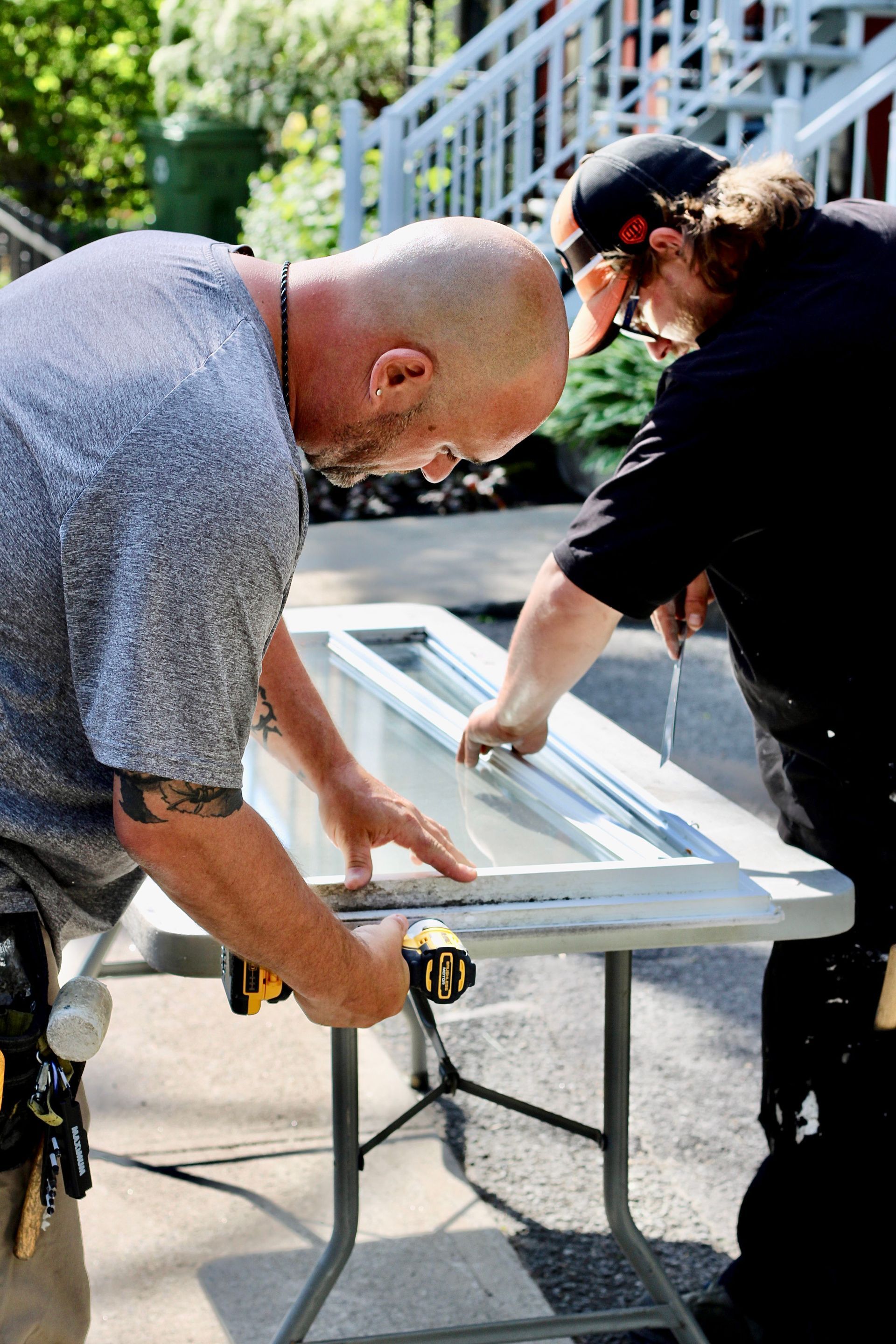 Two people assembling a white window frame on sawhorses outdoors