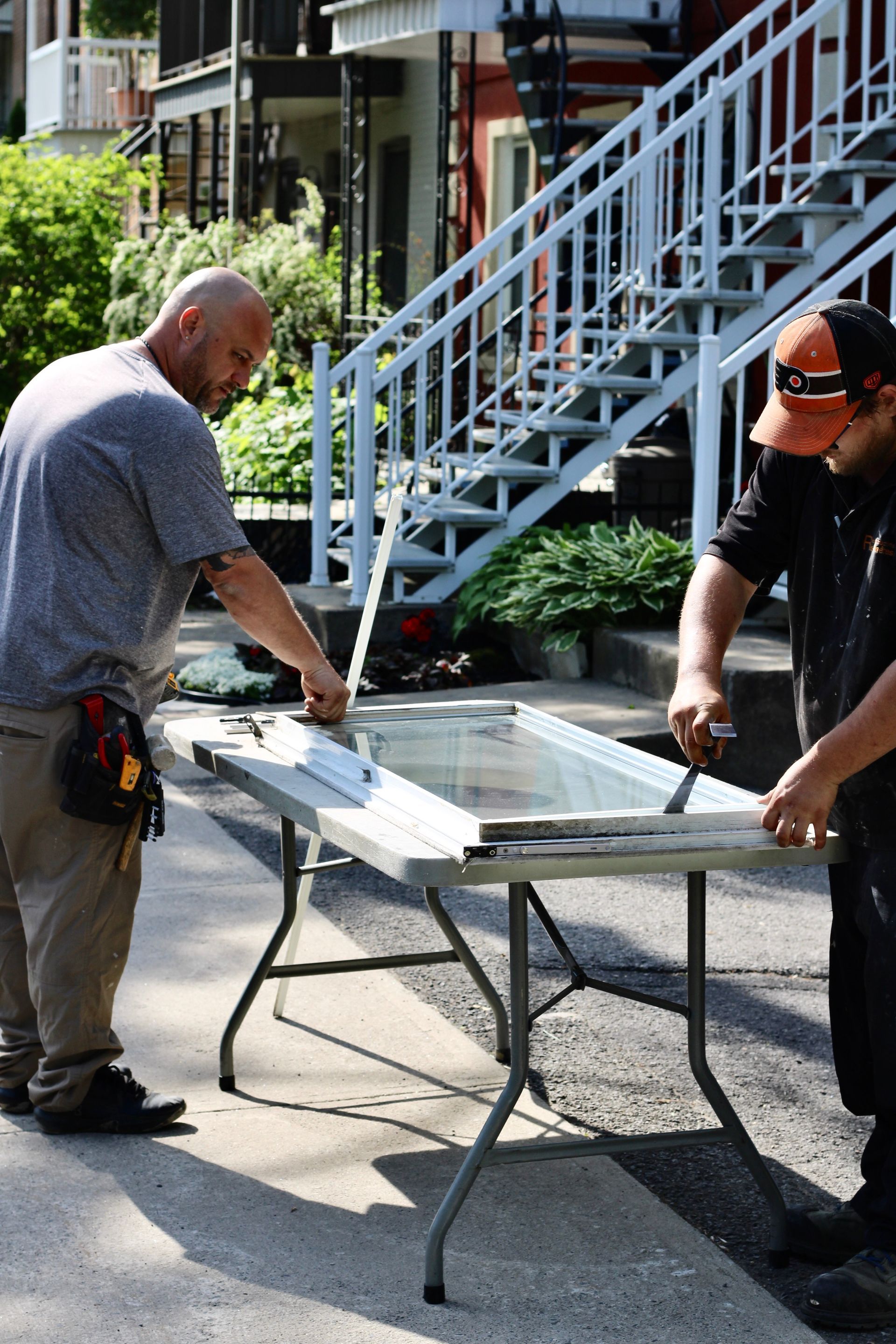 Two workers cutting a board on a folding table outside a building with stairs.
