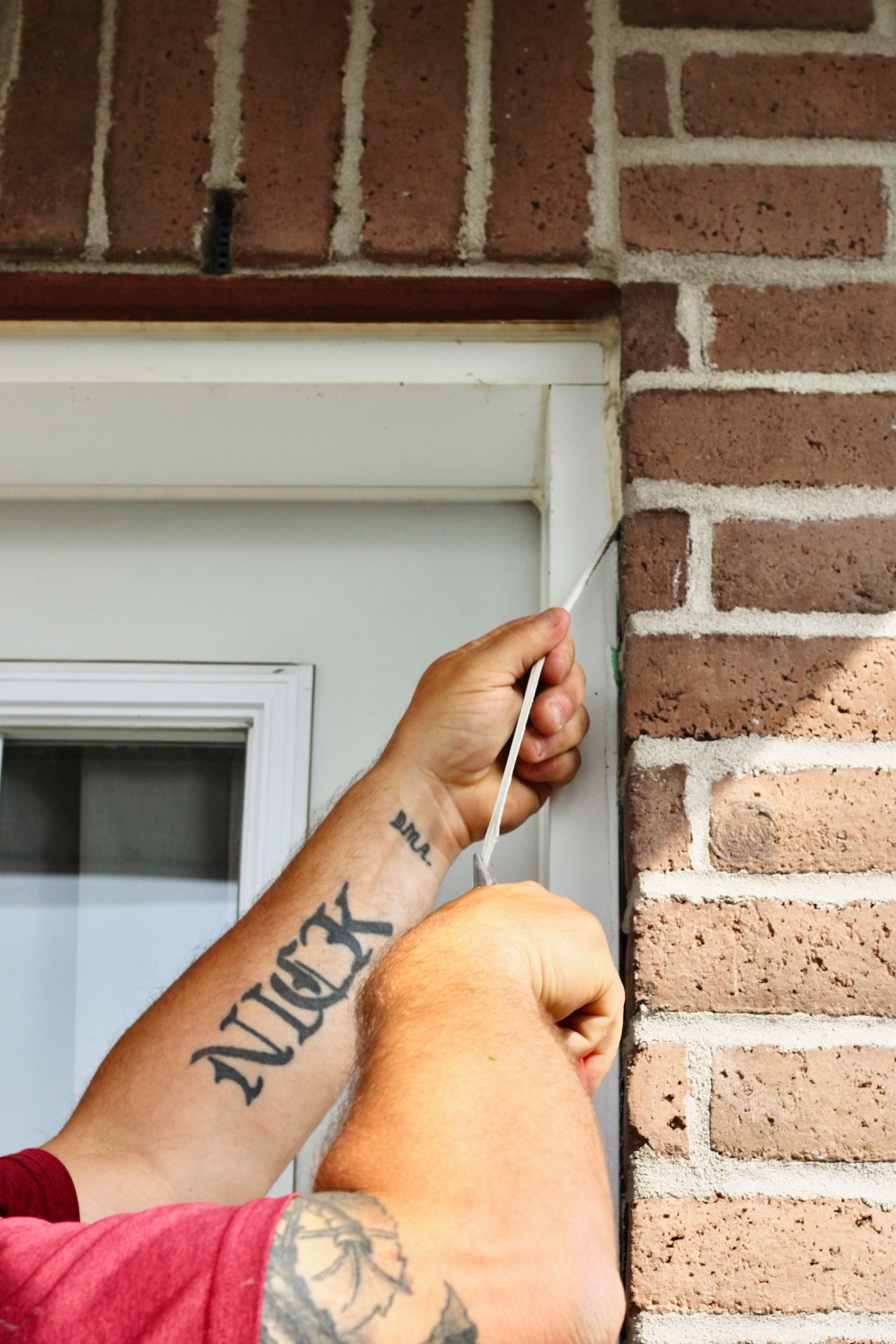 Hands caulk a white trim seam beside a brick wall above a window.