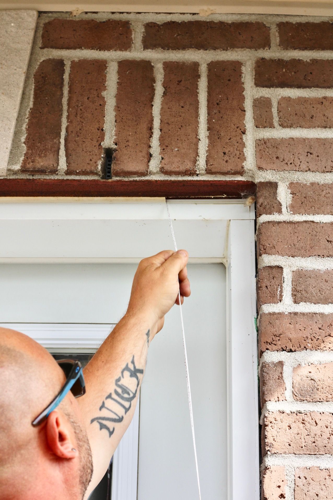 Hand measuring a white window frame beneath a brick wall with a tape measure
