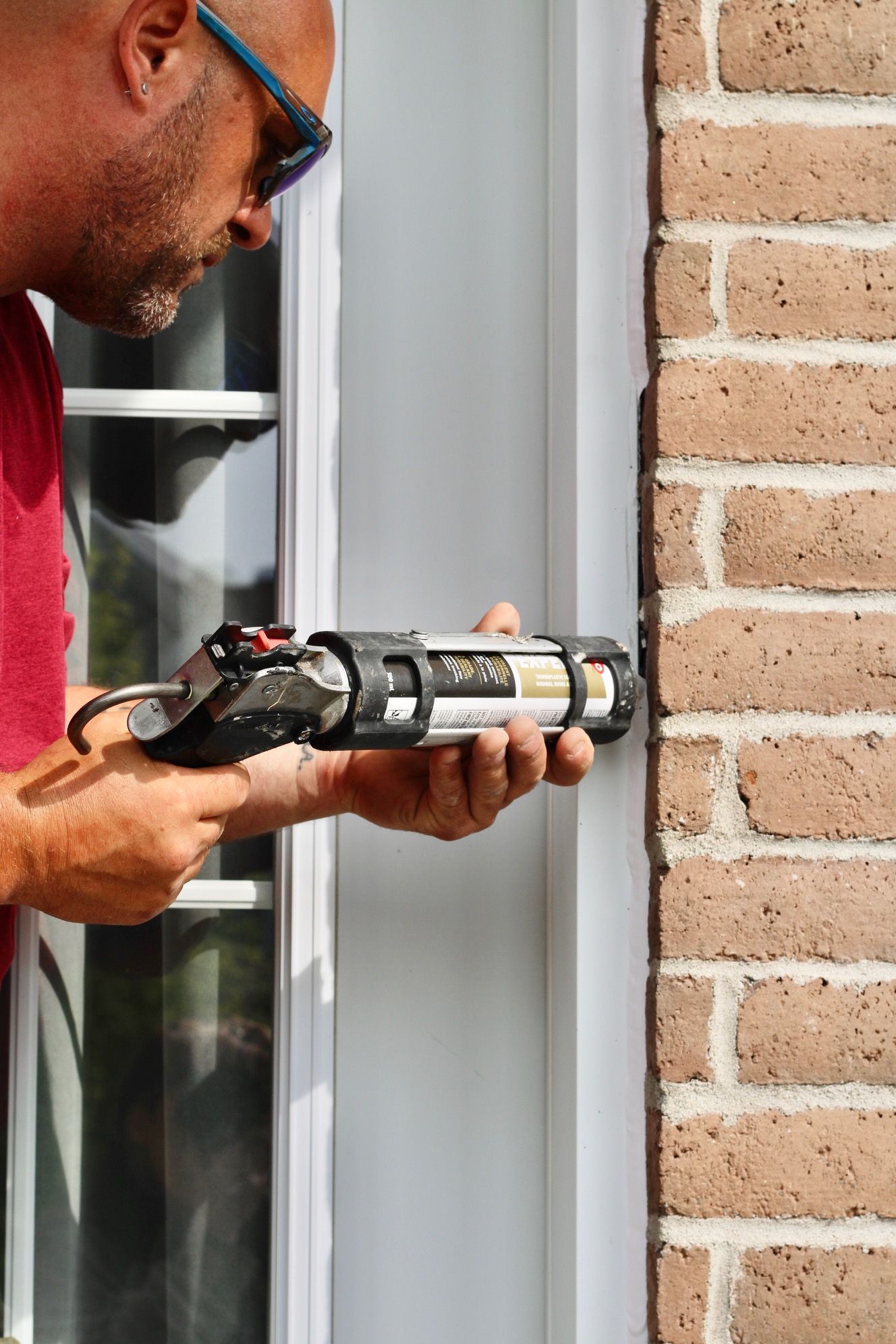 Person using a caulking gun to seal a window frame beside a brick wall