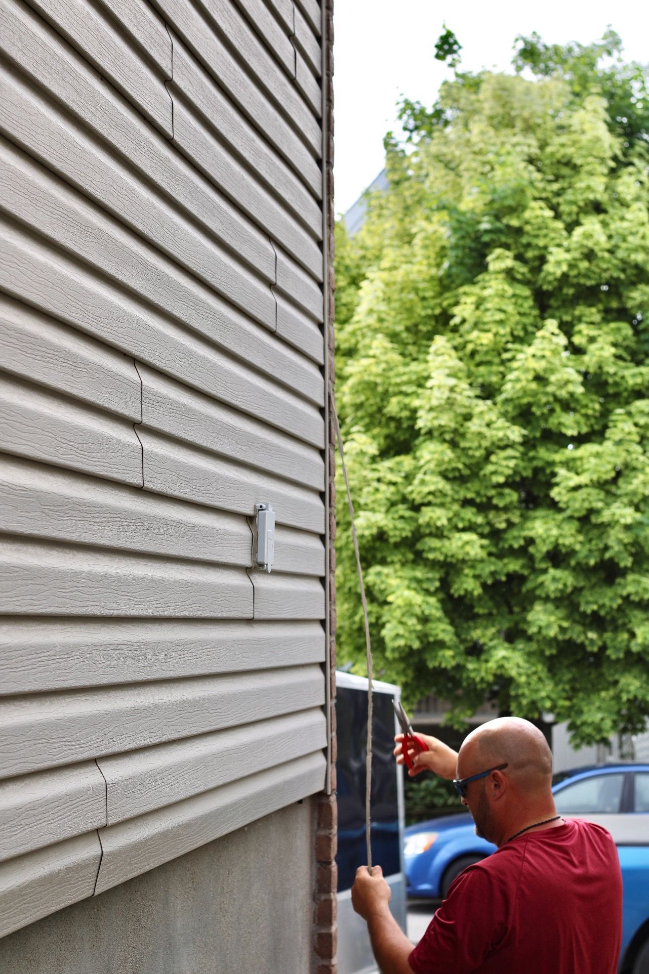 Person painting a house exterior with a roller beside beige siding and trees outdoors