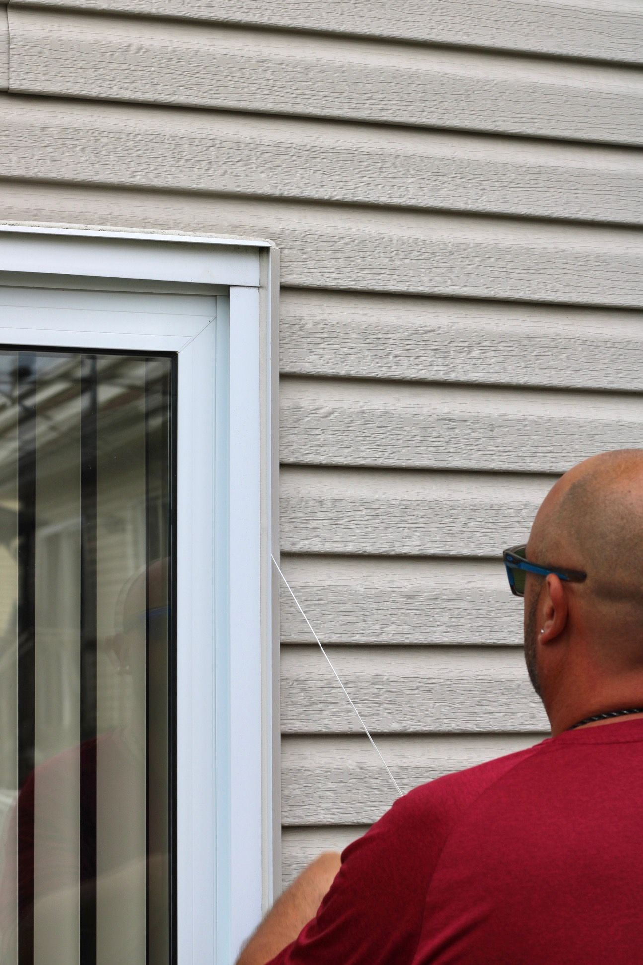 Person in a red shirt using a tool on tan house siding beside a white-framed window