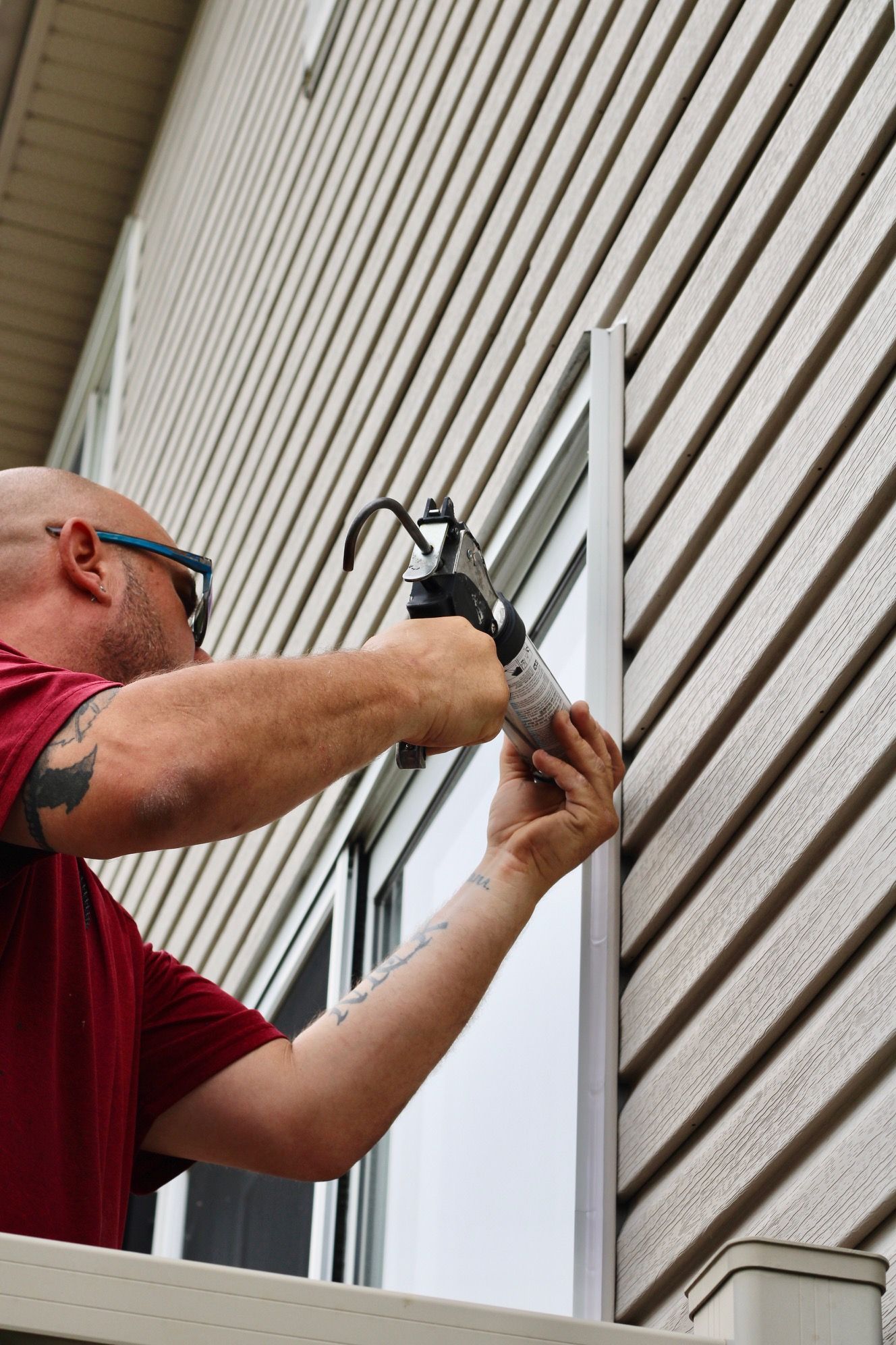 Worker caulking white vinyl siding beside a window on a house exterior