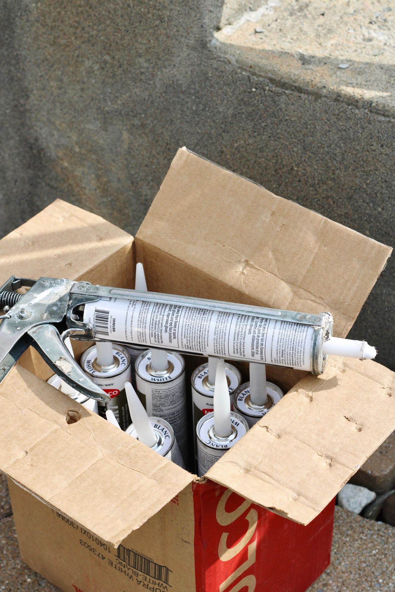 Open box of spray cans and a caulking gun on a concrete surface.