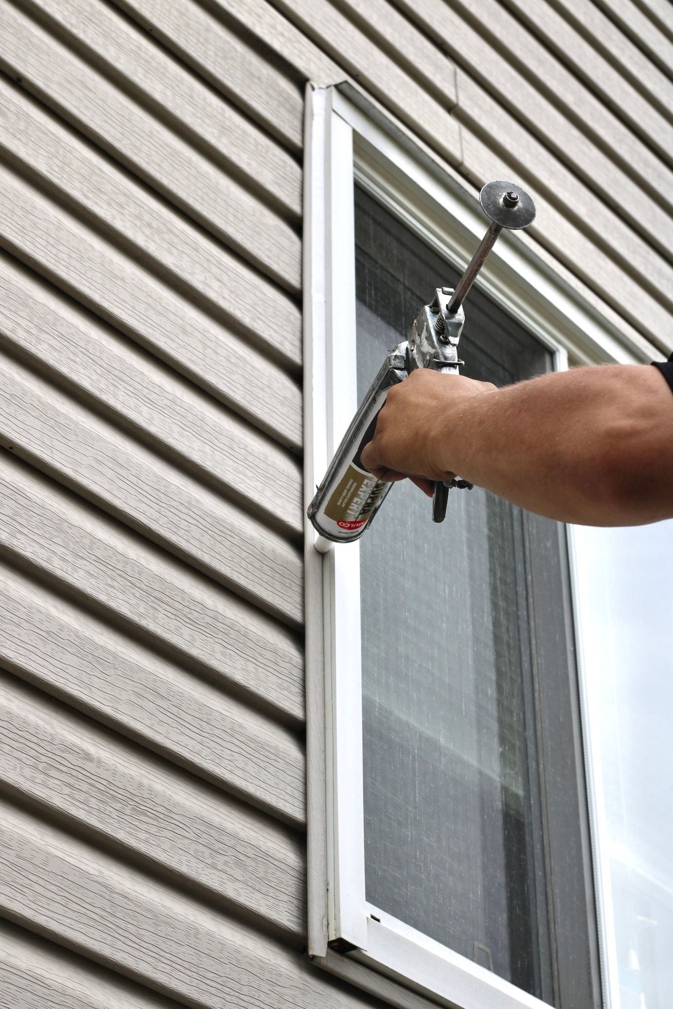 Person painting a window frame with a spray gun on a beige-sided house