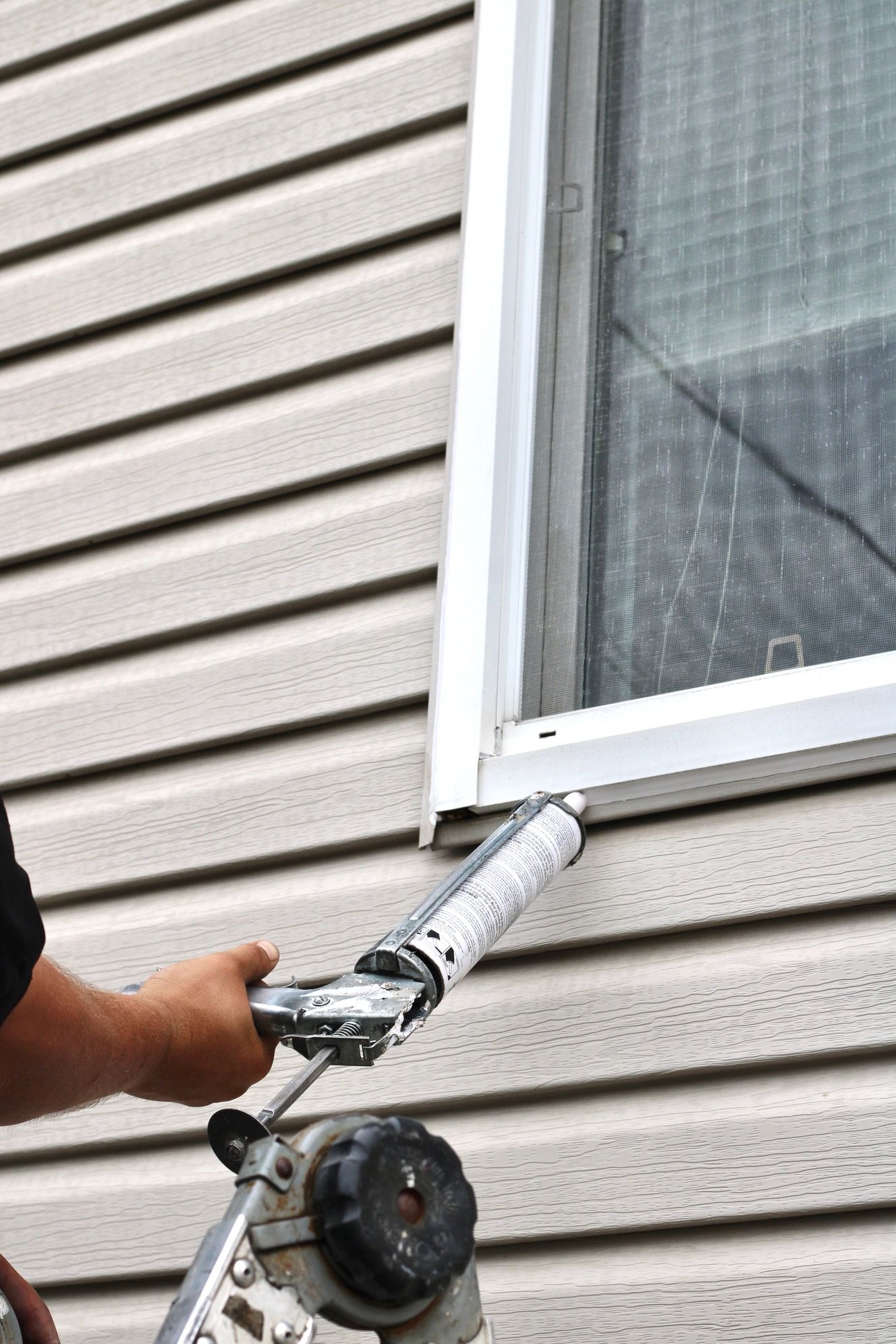 Worker applying caulk around a white window on beige vinyl siding
