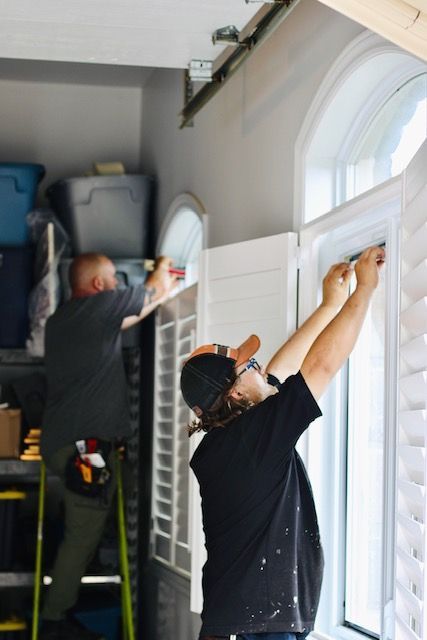 Two workers cleaning tall arched windows inside a bright room, using squeegees and tools.