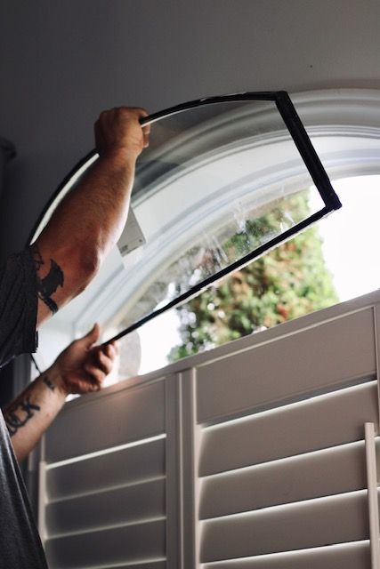 Person installing a black window screen inside a white window with shutters