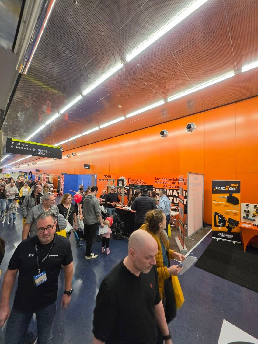 Crowded indoor event hall with orange walls, people walking and browsing booths under bright ceiling lights