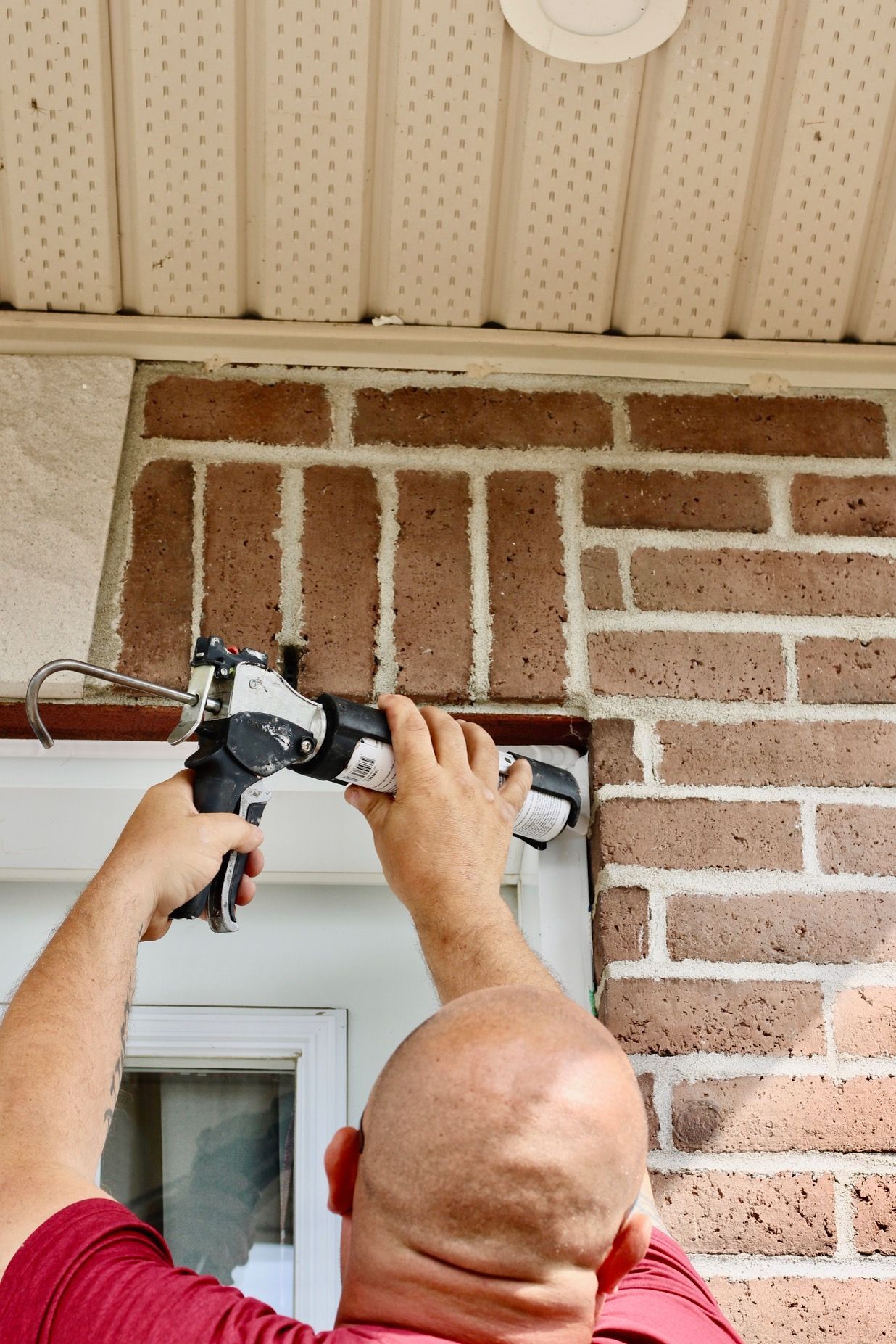 Person using a caulking gun to seal gaps above a window in a brick wall under a porch roof