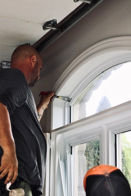 Person measuring trim around an arched window while standing on a ladder indoors