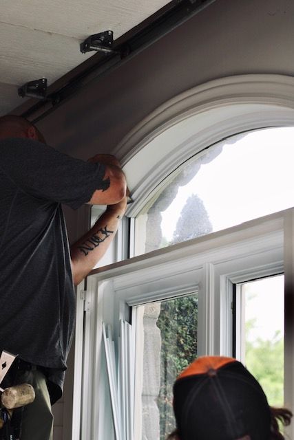 Worker installing trim around an arched window in a room with white walls and daylight.