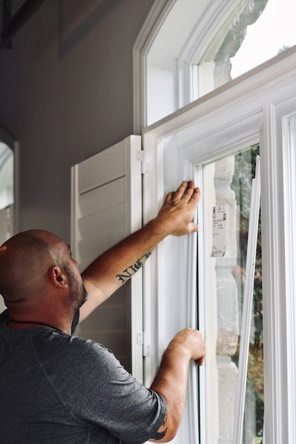 Man installing or adjusting a white window shutter beside a tall window in a bright room