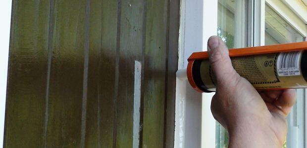 Hand applying orange caulk to a white window frame beside green-painted wood panels