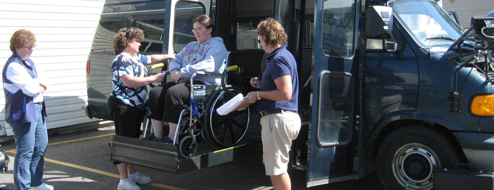 Two staff assist a person in a wheelchair onto a lift attached to a blue van.